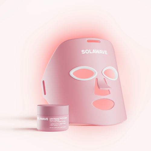 A pink LED face mask from Solawave, featuring cutouts for eyes, nose, and mouth, is displayed behind a matching pink jar labeled LightBoost Cream. The soothing background emphasizes its Red Light Therapy benefits that enhance skin radiance. This product is the Wrinkle Retreat Face Mask & LightBoost Cream Kit.